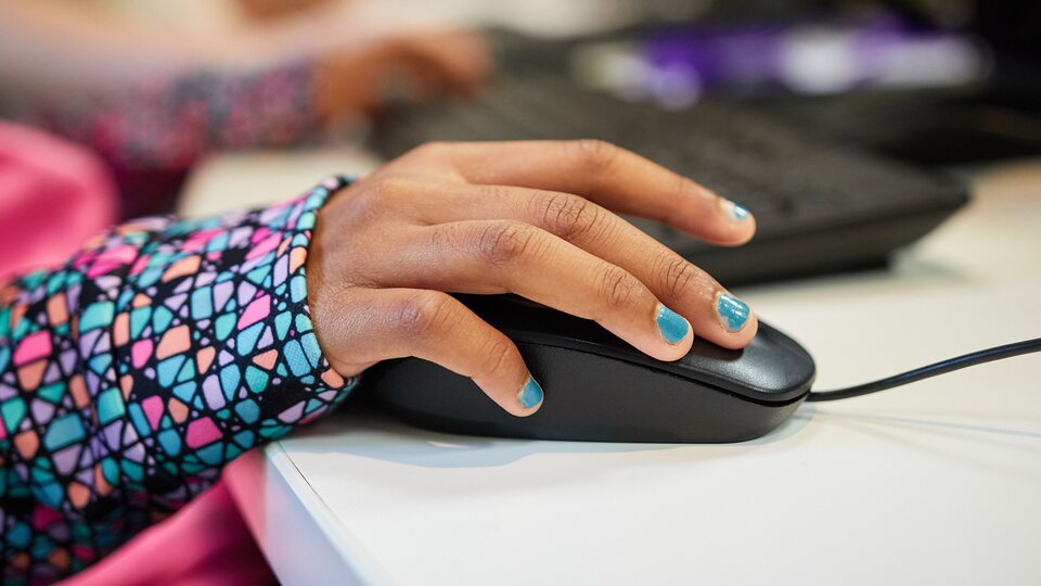 A student's hand resting on a mouse mid-lesson, with a classmate typing at the next desk